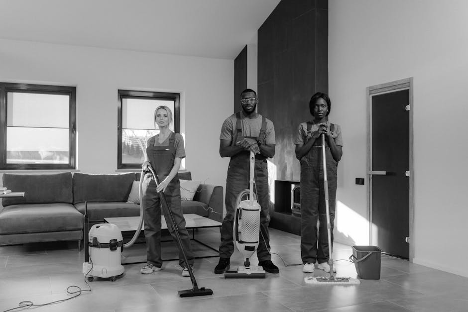 A team of three professional cleaners wearing matching dark uniforms are performing surface cleaning and deep cleaning in a modern, well-lit living room. The room features a large L-shaped sofa with a